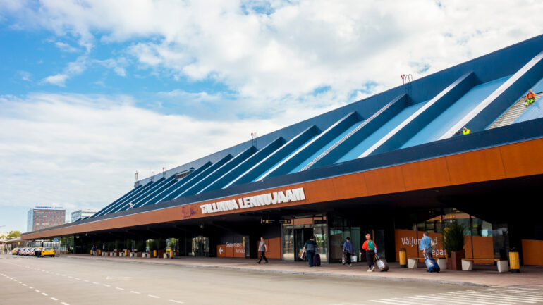 Fotol ehitusjärgus Tallinna Lennujaam. Hoone katus on sinist värvi, fassaad kollakaspruun. Hoone ees on neli inimest kohvritega lennujaama hoonesse sissepääsu suunas liikumas. Taamal on näha ootavaid taksosid.